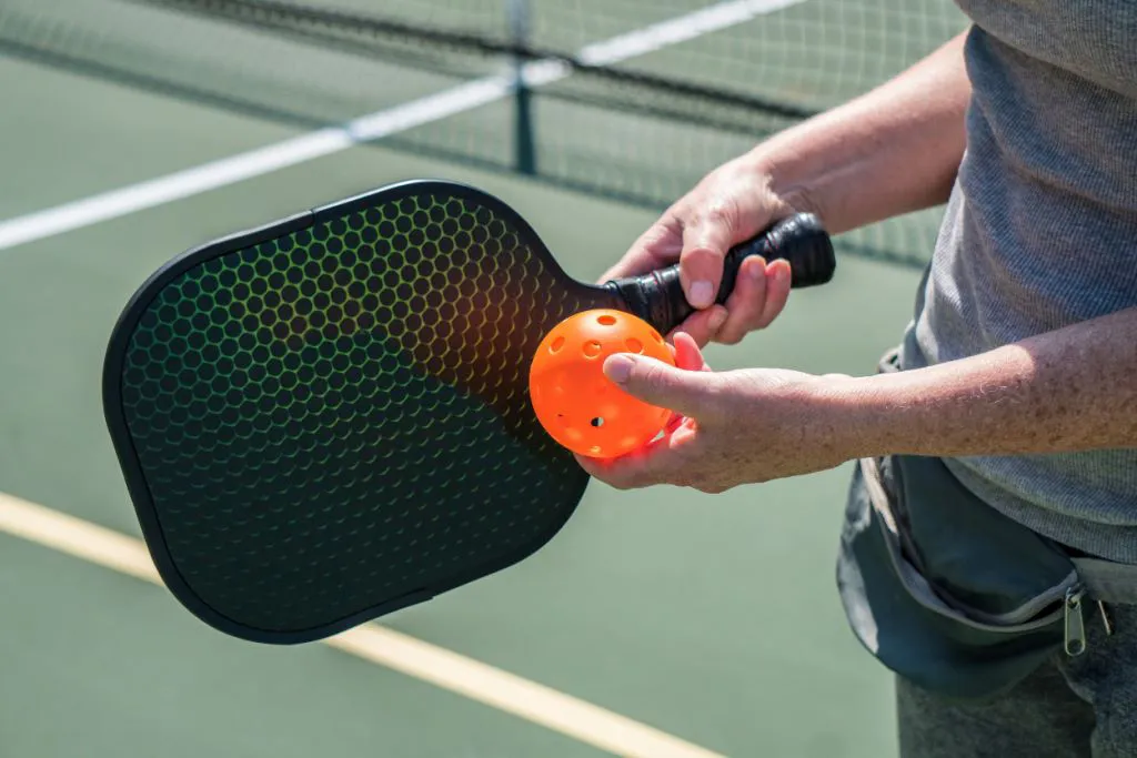 Hand holding a pickleball paddle and ball on a court.