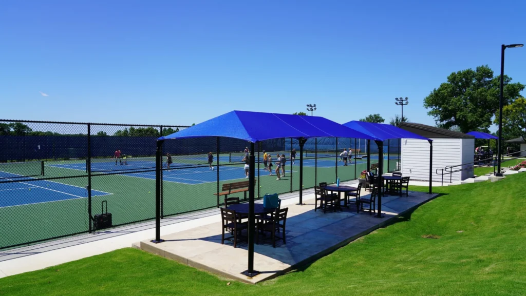Blue metal shade structure providing cover for benches near tennis courts.