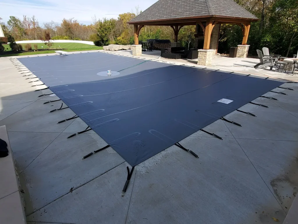 Gray mesh safety pool cover on stone deck – Pool with light gray fitted cover surrounded by stone paving.