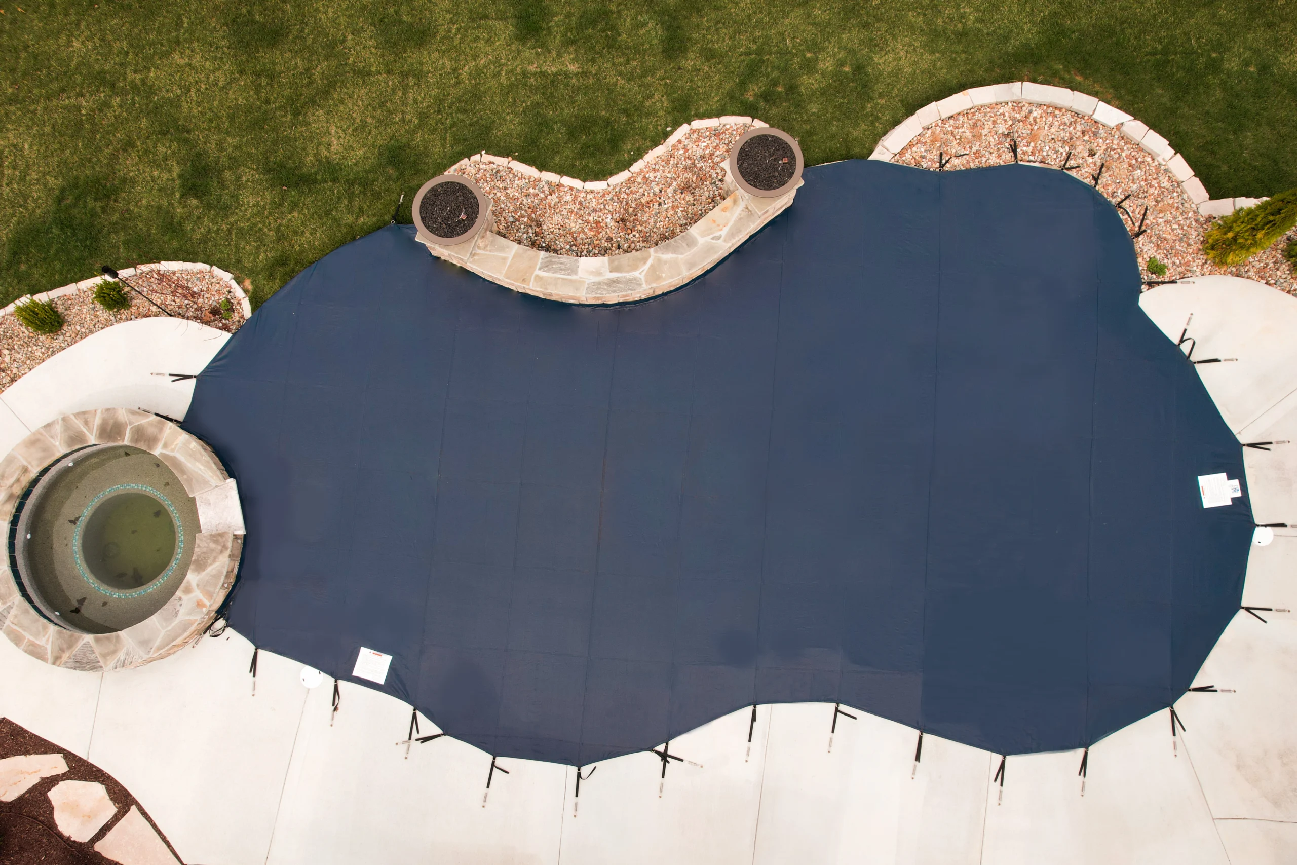 Dark blue custom-shaped pool safety cover fitted over in-ground backyard pool surrounded by stone patio
