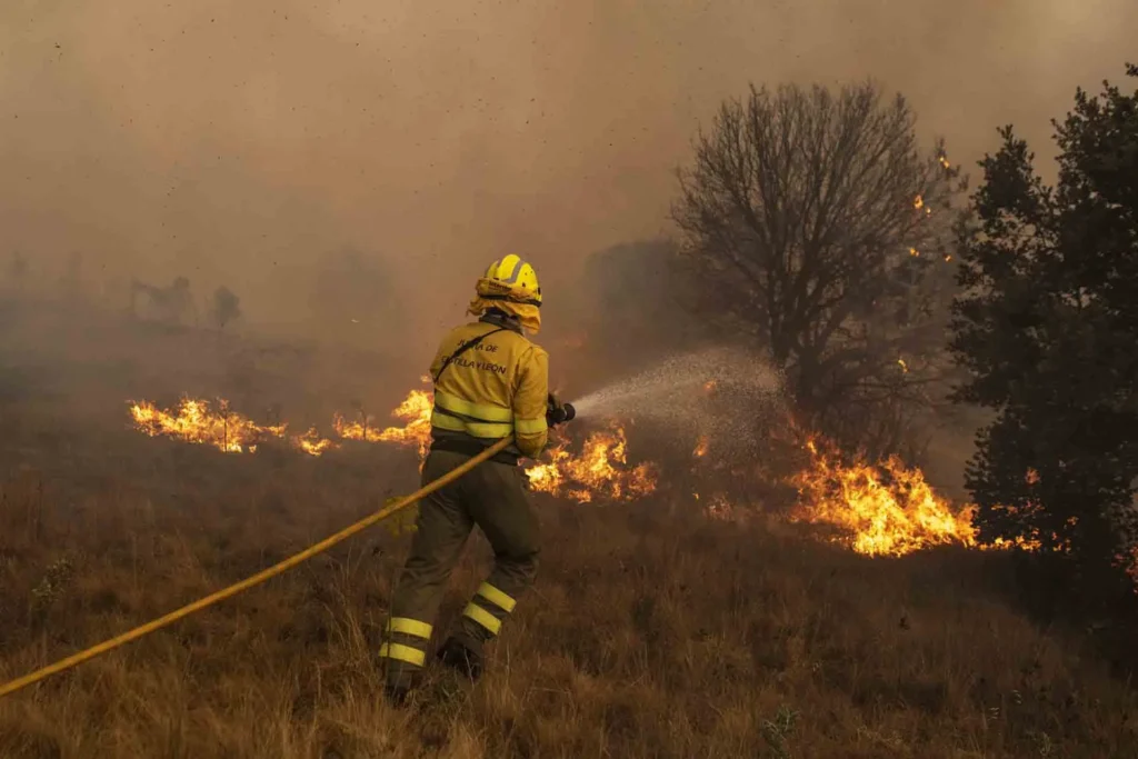 Firefighter in protective gear spraying water to control a grass fire in an open field.