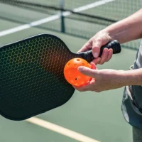Hand holding a pickleball paddle and ball on a court.